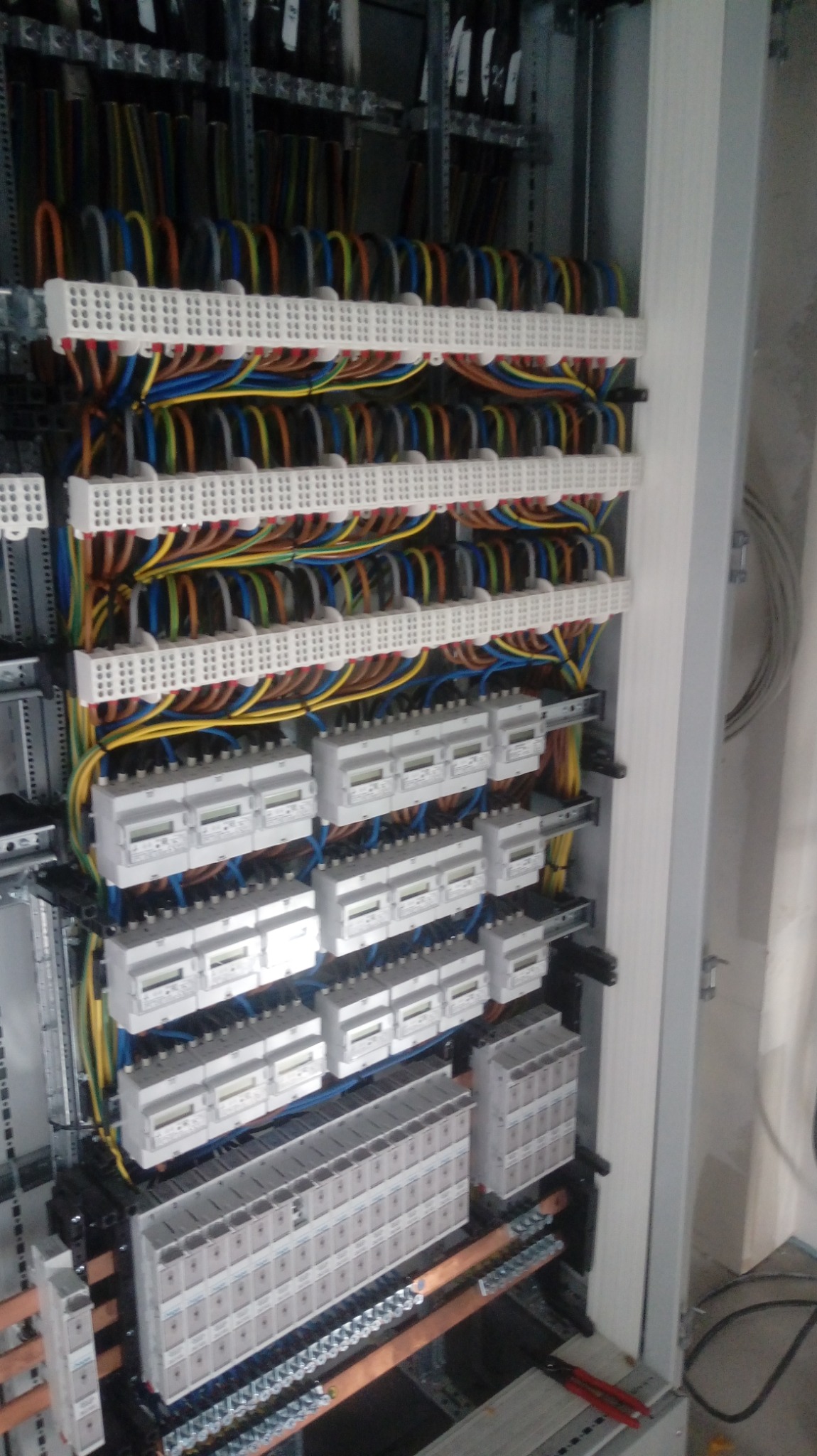 Rozdzielnica elektryczna z licznymi przewodami w różnych kolorach, listwami zaciskowymi i modułowymi licznikami energii elektrycznej, zamontowana w metalowej szafie, widoczne narzędzia.