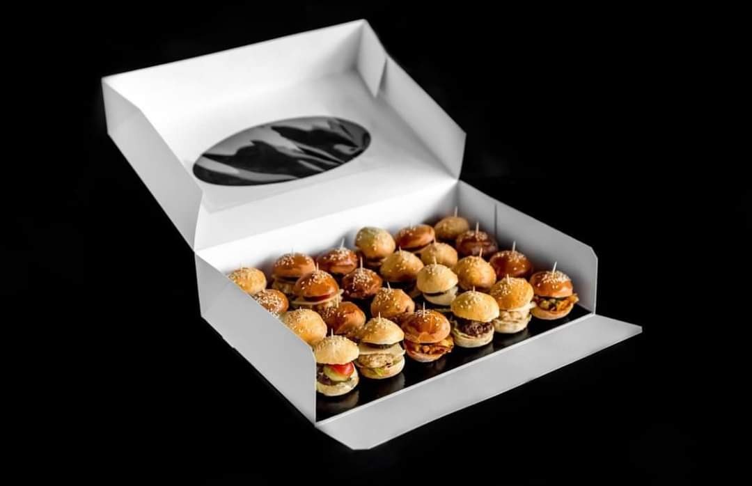 Otwarty biały karton z okienkiem wypełniony rzędami mini burgerów na czarnym tle, każdy burger z innym nadzieniem, spięty wykałaczką.