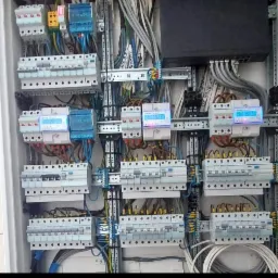 Rozdzielnia elektryczna z zamontowanymi bezpiecznikami, licznikami energii z wyświetlaczami wskazującymi zużycie, oraz licznymi przewodami w kolorach szarym, białym, niebieskim i żółto-zielonym...