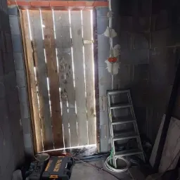 Surowy remont łazienki z drewnianymi drzwiami z prześwitami, widocznymi kablami elektrycznymi na ścianie z szarych bloków, drabina i skrzynki narzędziowe DeWalt na podłodze w trakcie prac.