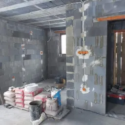 Surowy stan wnętrza w trakcie budowy: ściany z szarych bloczków, widoczne przewody elektryczne w ścianach, puszki instalacyjne, worki z zaprawą murarską i wiadro na podłodze, surowy betonowy strop.