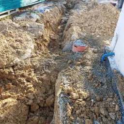 Wykop w ziemi obok budynku z widoczną rurą kanalizacyjną i stosem desek w tle.