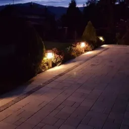 Oświetlony ścieżką z kostki brukowej w ogrodzie po zmierzchu, z lampkami wzdłuż krawędzi, widok na zalesione wzgórza w tle.