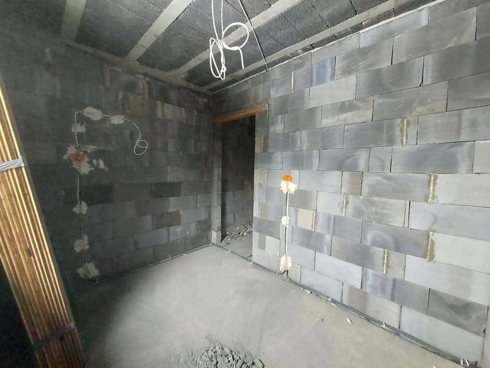 Surowy stan wnętrza z szarych bloczków betonowych, widoczne przewody elektryczne wystające ze ściany, betonowa wylewka podłogowa i drewniane listwy przypodłogowe w rogu.