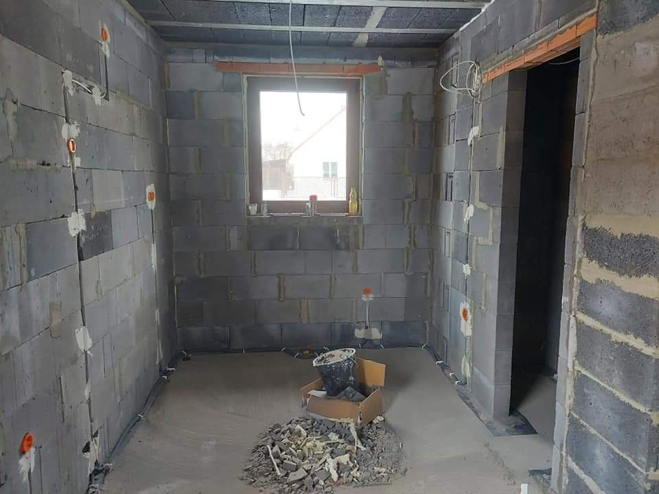 Surowy stan łazienki w trakcie remontu: ściany z bloczków, widoczne instalacje elektryczne w peszlach, puszki pod gniazdka, okno z parapetem, gruz na podłodze, przygotowanie do dalszych prac...