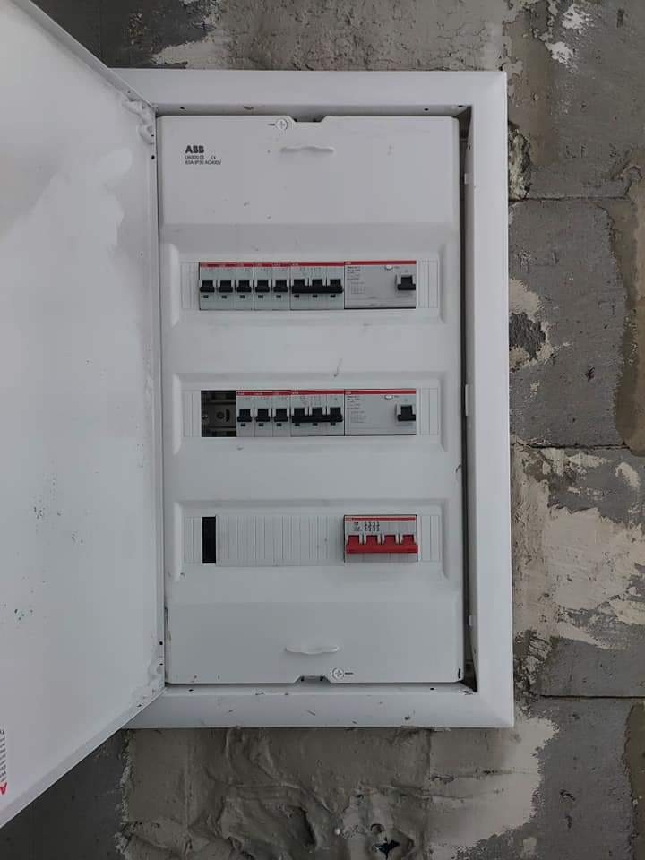 Otwarta, biała skrzynka elektryczna ABB z trzema rzędami bezpieczników na tle surowej, szarej ściany z cegły.