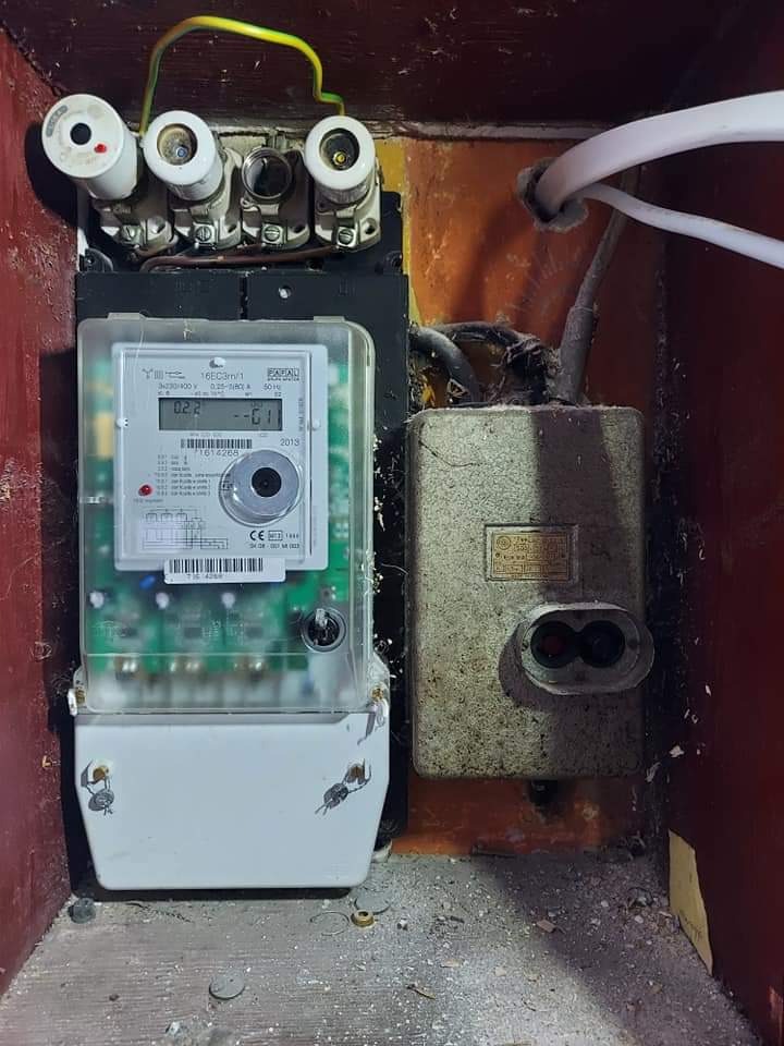 Stary licznik elektryczny z bezpiecznikami topikowymi i wyłącznikiem, widoczne ślady użytkowania i zabrudzenia.