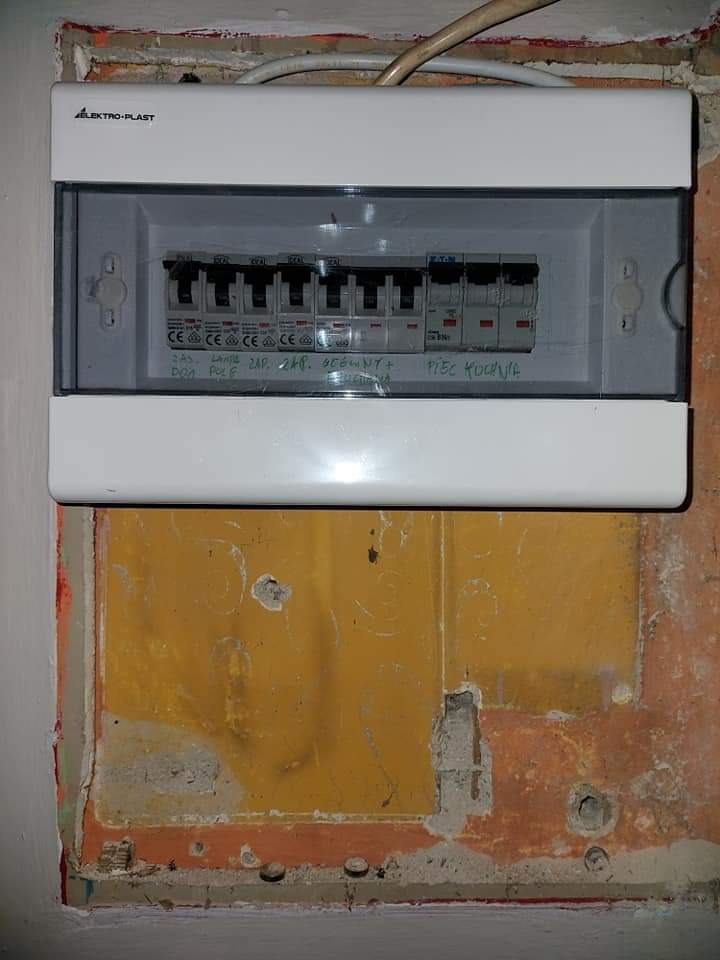 Biała skrzynka elektryczna z bezpiecznikami marki Elektro-Plast zamontowana na pomalowanej na żółto ścianie z ubytkami tynku i widocznymi śladami po instalacji.