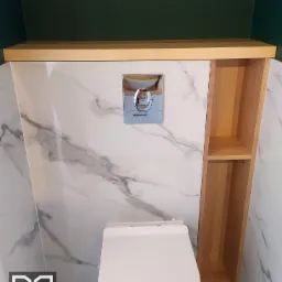 Nowoczesna toaleta podwieszana z zabudową z jasnego drewna i marmurowymi płytkami na ścianach, widoczny chromowany przycisk spłuczki i logo firmy meblarskiej w lewym dolnym rogu.