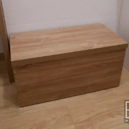 Drewniana skrzynia z siedziskiem w odcieniach brązu, umieszczona w rogu łazienki z jasnymi płytkami i podłogą imitującą drewno, widoczne logo producenta w prawym dolnym rogu.