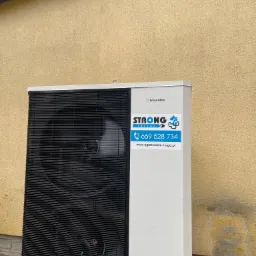 Zewnętrzna jednostka pompy ciepła Hewalex z naklejką firmy instalacyjnej Strong Instal na tle otynkowanej ściany budynku.