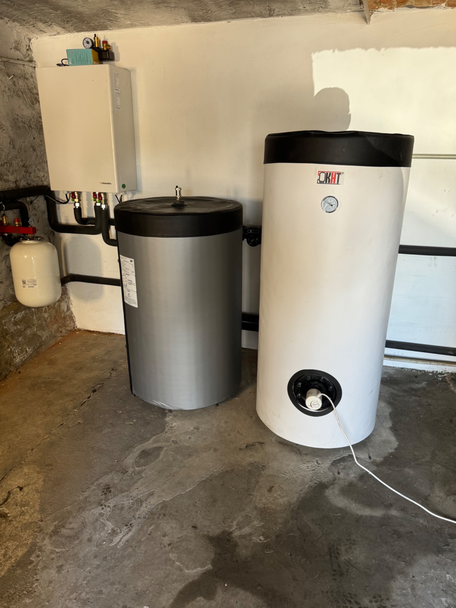 Nowoczesny system grzewczy z dwoma zbiornikami akumulacyjnymi (szary i biały) oraz wiszącym piecem grzewczym na tle surowej ściany i betonowej podłogi.