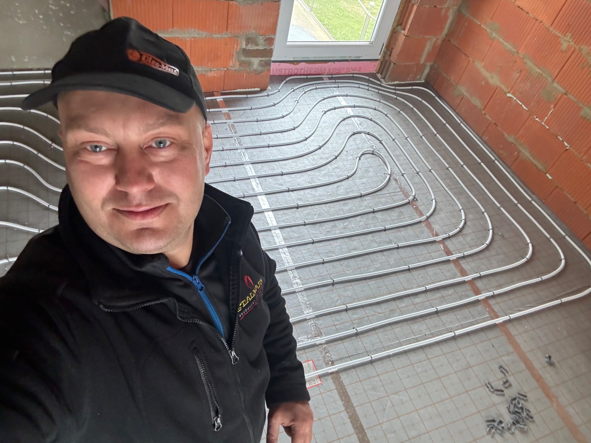 Instalator w czarnej kurtce i czapce z daszkiem pozuje na tle rozłożonych rur ogrzewania podłogowego w surowym stanie, widoczne mocowania rur i ceglany mur w tle.