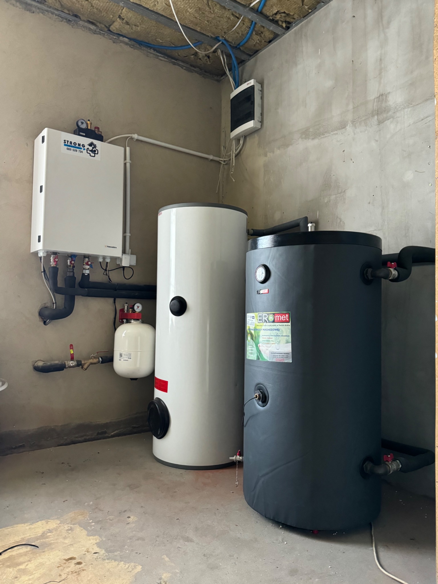 Zainstalowany system grzewczy z białym piecem i dwoma zbiornikami akumulacyjnymi: białym i grafitowym, w surowym pomieszczeniu z widoczną instalacją elektryczną.