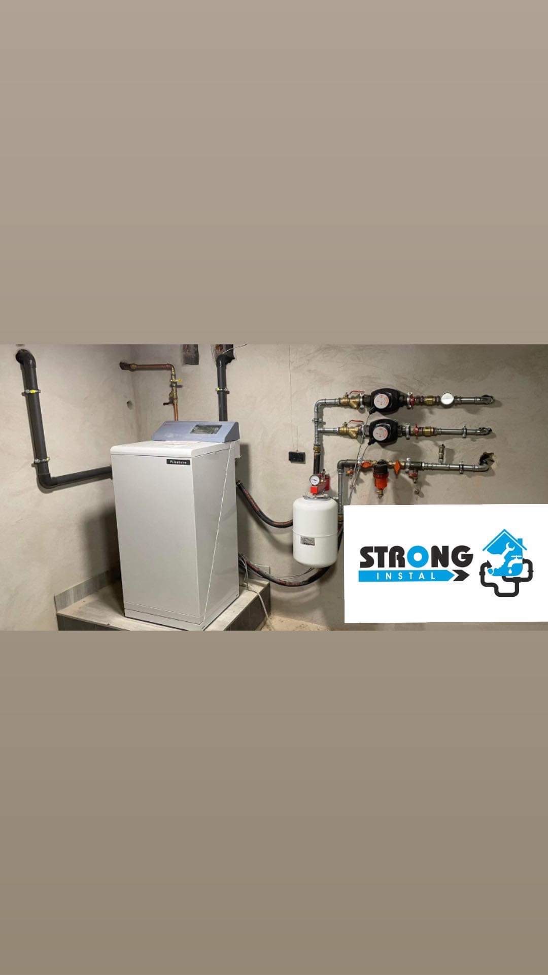 Biały piec grzewczy z szarym panelem sterowania i podłączoną instalacją hydrauliczną w surowym wnętrzu, obok widoczne logo firmy instalacyjnej.