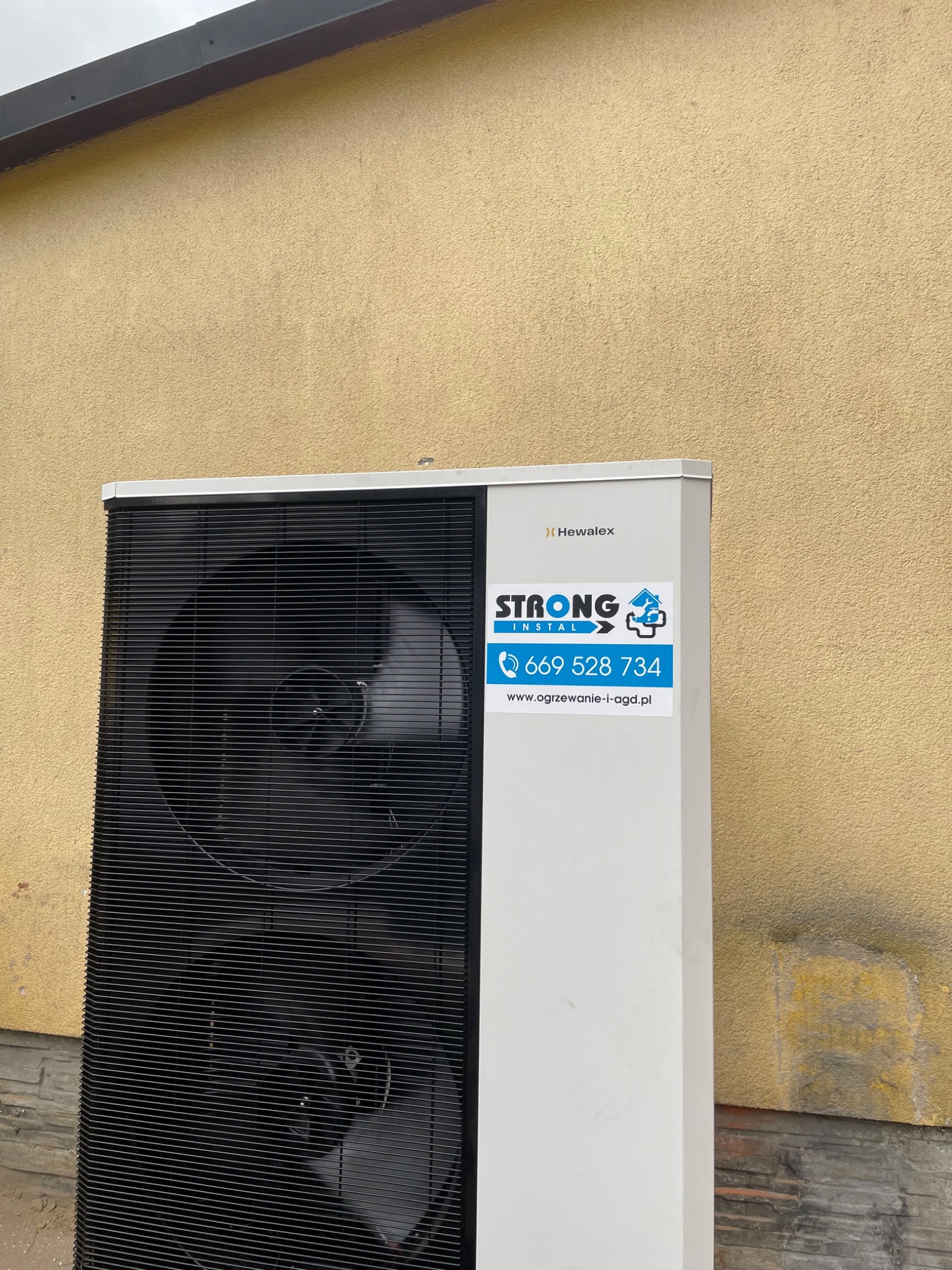 Zewnętrzna jednostka pompy ciepła Hewalex z naklejką firmy instalacyjnej Strong Instal na tle otynkowanej ściany budynku.