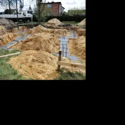 Wykopane fundamenty domu z częściowo wylanym betonem i wystającymi prętami zbrojeniowymi, na pierwszym planie trawa, w tle budynki mieszkalne i pompa do betonu.