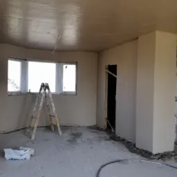Pokój po nałożeniu gładzi gipsowej na ściany i sufit, widoczna drabina malarska, okno z zaokrąglonym parapetem i resztki materiałów budowlanych na podłodze.