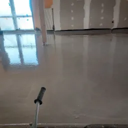 Wylewka samopoziomująca w trakcie rozprowadzania, widoczne narzędzie z uchwytem oraz fragment okna zabezpieczonego folią malarską.