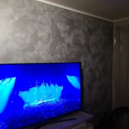 Dekoracyjna ściana w odcieniach szarości z efektem przetarć, widoczny fragment telewizora z jaskrawym obrazem koncertu.