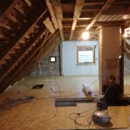 Poddasze w trakcie adaptacji: widoczne drewniane krokwie, ocieplona ściana szczytowa z oknem, płyty OSB na podłodze, zwój materiału izolacyjnego i narzędzia, instalacja elektryczna w trakcie montażu.