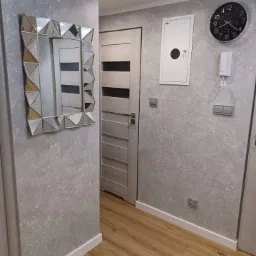 Korytarz po remoncie z szarą, teksturowaną ścianą, lustrem w geometrycznej ramie, drewnopodobną podłogą, drzwiami wewnętrznymi z poziomymi panelami, zegarem ściennym i białą skrzynką elektryczną.