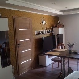 Wagner Tomasz - Nowoczesny salon z jadalnią, wyróżniający się teksturowaną, złotobrązową ścianą, drewnianymi drzwiami z poziomymi wstawkami i podwieszanym sufitem z oświetleniem LED.