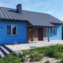 Wagner Tomasz - Nowoczesny dom z jaskrawoniebieską elewacją, ciemnoszarym dachem z blachy na rąbek i drewnianymi akcentami. Przed nim wybrukowany taras z szarych kostek, otoczony zielenią.