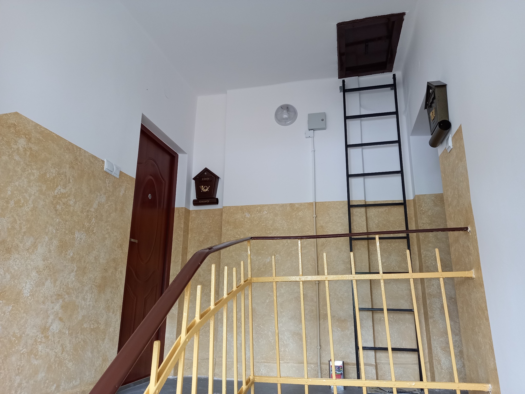 Klatka schodowa po odświeżeniu, ściany pomalowane na biało i beżowo z efektem dekoracyjnym, żółta metalowa balustrada, brązowe drzwi wejściowe i drabina do włazu na strych.