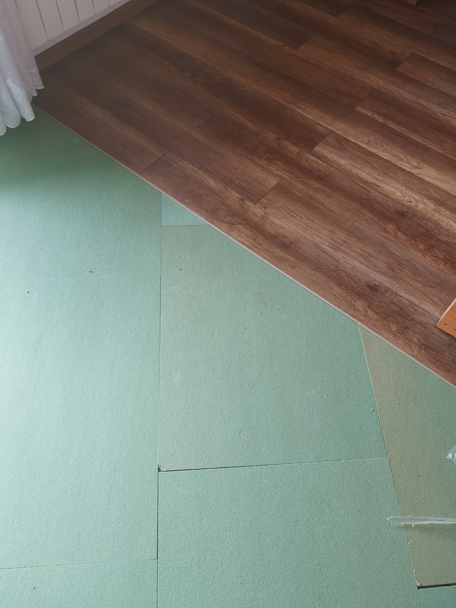 Fragment podłogi z brązowymi panelami ułożonymi częściowo na zielonym podkładzie izolacyjnym.