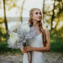 Panna młoda w białej sukni trzyma bukiet z białych hortensji i gipsówki na tle zieleni.