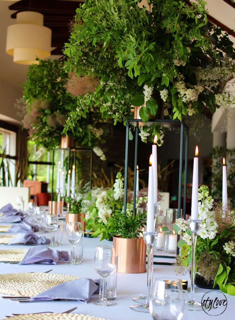 Dekoracja stołu weselnego z wysokimi świecami w srebrnych świecznikach, kompozycjami kwiatowymi w geometrycznych, metalowych konstrukcjach i miedzianych donicach, oraz fioletowymi serwetkami...