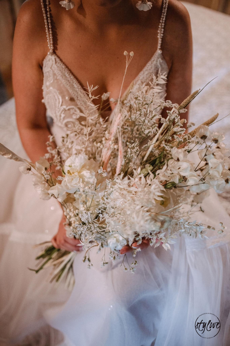 Panna młoda w koronkowej sukni trzyma bukiet ślubny z białych kwiatów i suszonych traw, widoczne delikatne kolczyki.