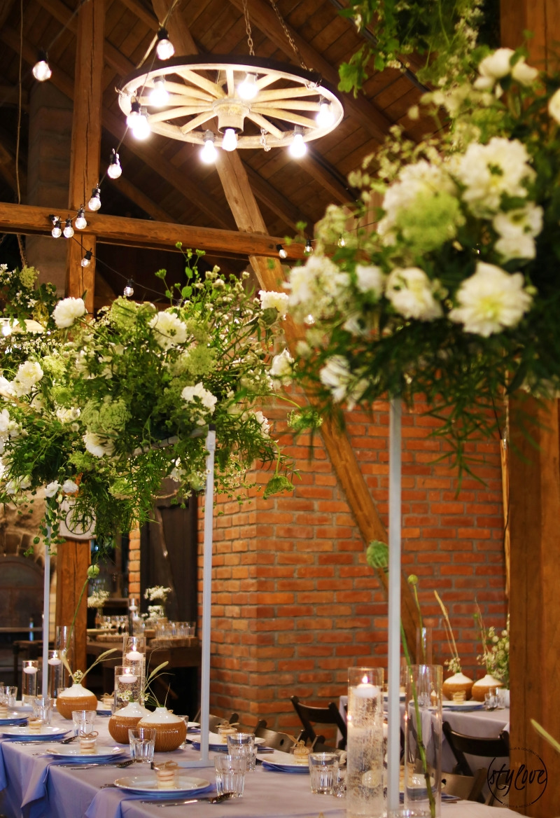 Dekoracja sali weselnej z drewnianą konstrukcją dachu, oświetleniem w stylu wozu i wysokimi kompozycjami kwiatowymi na stołach.
