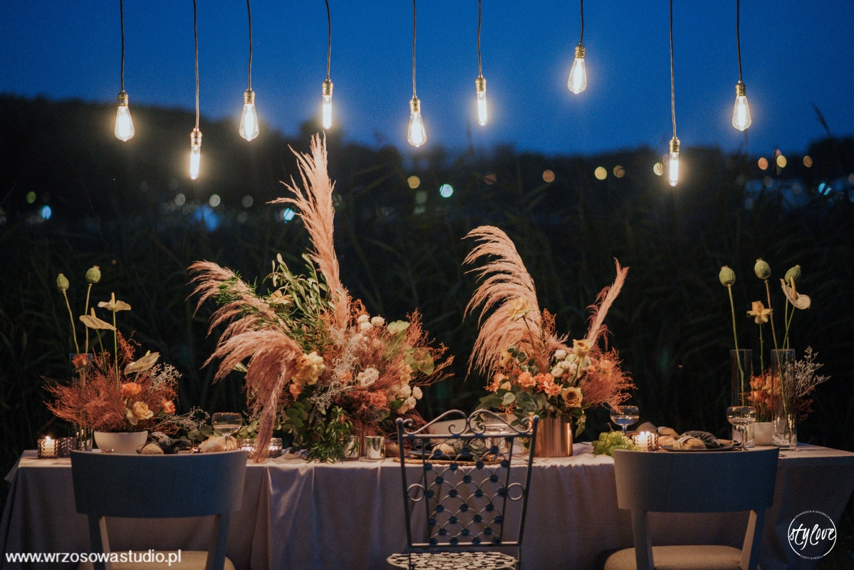 Uroczysta dekoracja stołu weselnego z kompozycjami kwiatowymi z pampasów, oświetlona wiszącymi żarówkami w stylu retro, widok na zewnątrz w tle.