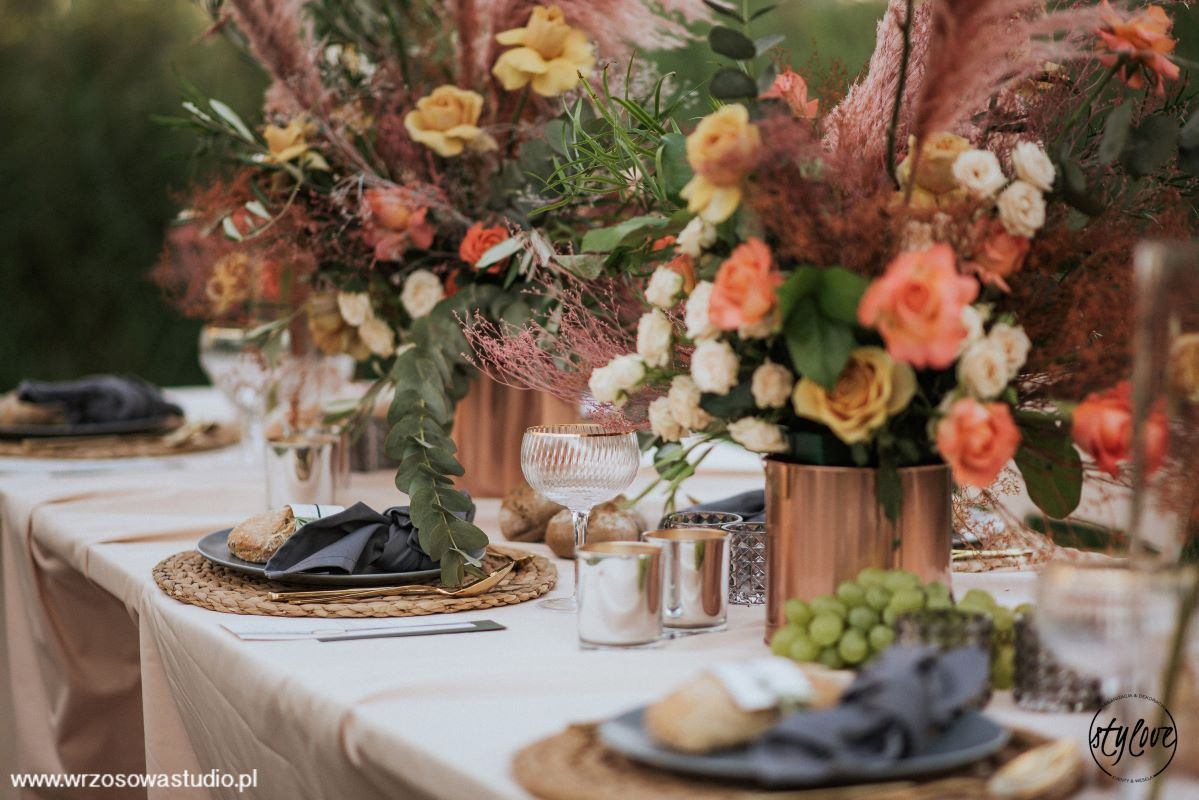 Dekoracja stołu weselnego z kompozycjami kwiatowymi w miedzianych wazonach, szarymi serwetkami i złotymi sztućcami na plecionych podkładkach.
