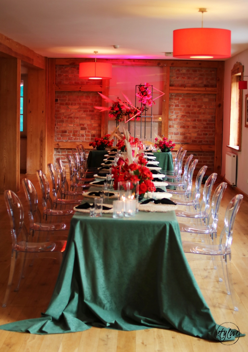 Uroczysty stół z zielonym obrusem, nakryty białymi talerzami z czarnymi serwetkami, ozdobiony kompozycjami z czerwonych róż i świecami, oświetlony czerwonymi lampami wiszącymi