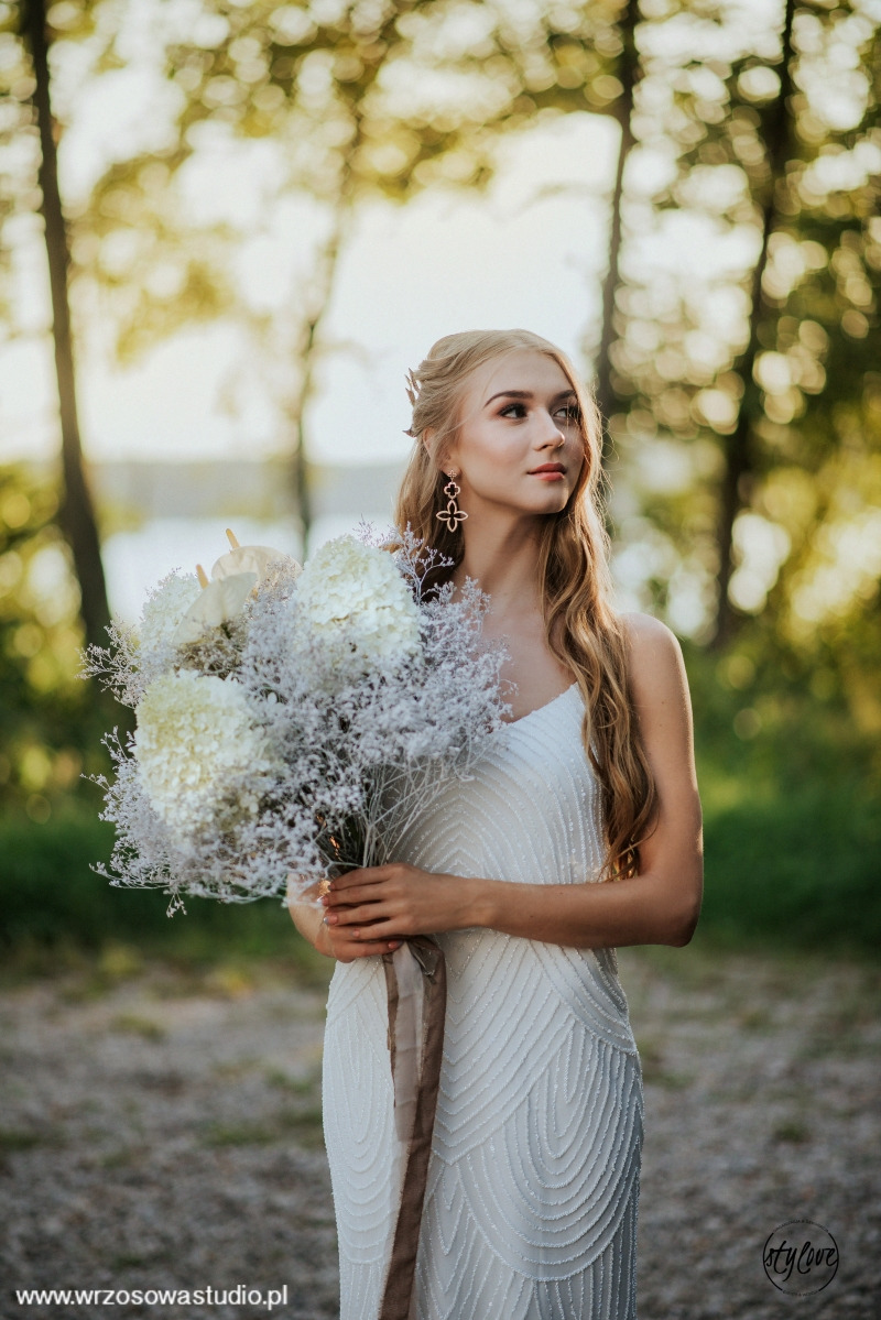 Panna młoda w białej sukni trzyma bukiet z białych hortensji i gipsówki na tle zieleni.