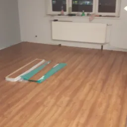 Brązowe panele podłogowe ułożone w pustym pokoju, z oknem w tle i zapakowanymi listwami przypodłogowymi oraz panelami czekającymi na montaż.