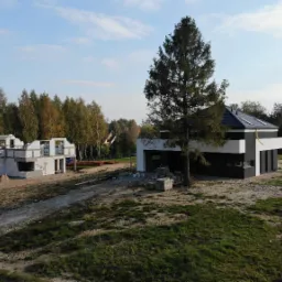 Dwa budynki w różnym stadium budowy: jeden w stanie surowym otwartym z widocznymi bloczkami, drugi z elewacją w kolorach czerni i bieli, otoczone zielenią i drzewami.