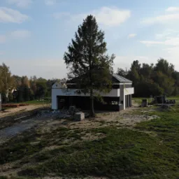 Dwa domy w różnym stadium budowy: jeden w stanie surowym, drugi z ocieploną, biało-czarną elewacją i ukończonym dachem, na pagórkowatym terenie z drzewami w tle.