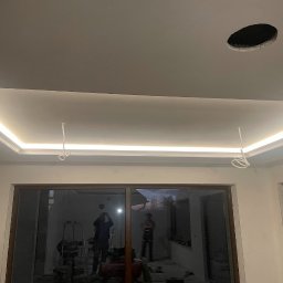 Roksz Michał Usługi Ogólnobudowlane - Podwieszany sufit z oświetleniem LED w narożach, widoczne kable i otwór na lampę. W tle ekipa remontowa i okno.