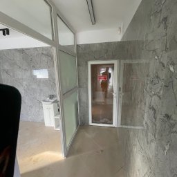 Roksz Michał Usługi Ogólnobudowlane - Wykończona łazienka z marmurowymi płytkami, białą umywalką i szklanymi drzwiami. Widoczne detale wykończeniowe i oświetlenie.