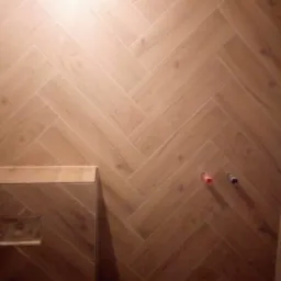 Ściana wyłożona płytkami imitującymi drewno w układzie jodełki, widoczne przyłącza wodne w kolorze czerwonym i niebieskim oraz fragment zabudowy geberit