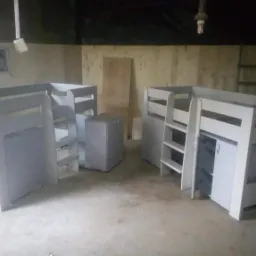 Dwa białe łóżka piętrowe z drabinkami i szafkami, ustawione w warsztacie z surowymi ścianami i podłogą betonową, widoczna drabina oparta o ścianę w tle.