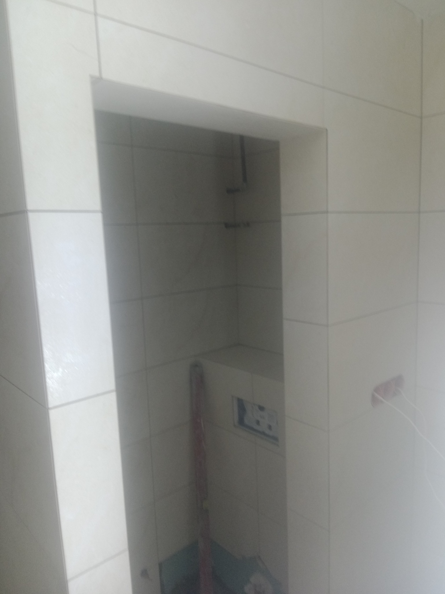 Narożnik w łazience z wykończonymi jasnymi, kwadratowymi płytkami ceramicznymi, widoczne otwory na instalacje i fragment pionowego elementu instalacyjnego w niszy.