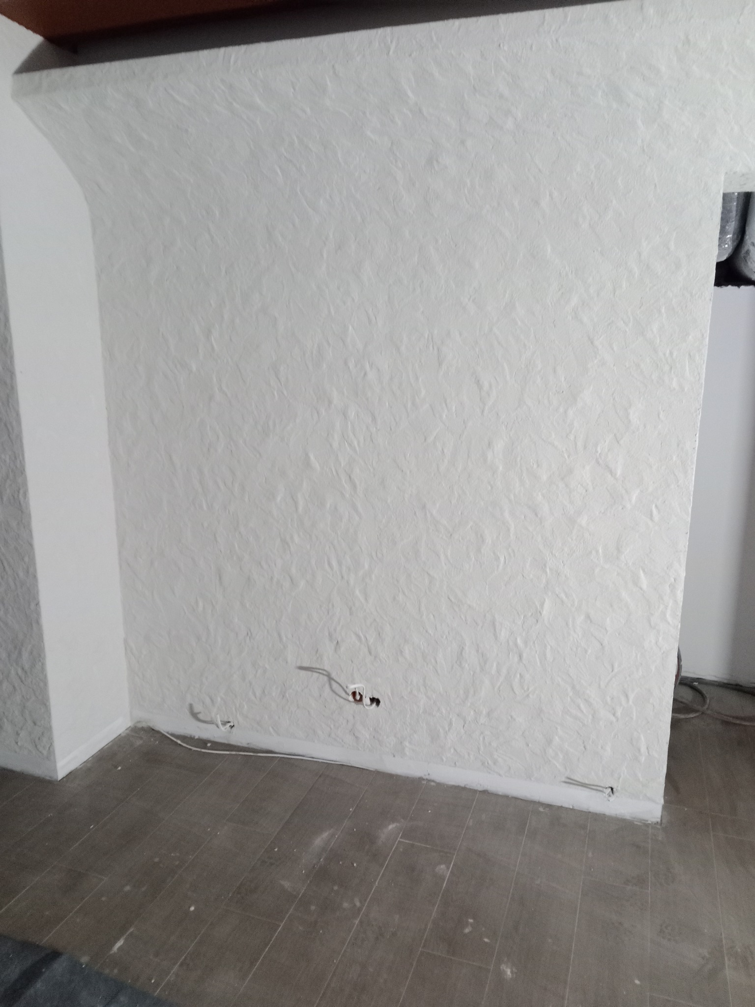 Ściana z dekoracyjną, białą strukturą, widoczne gniazdka elektryczne i fragment podłogi z płytek imitujących drewno.