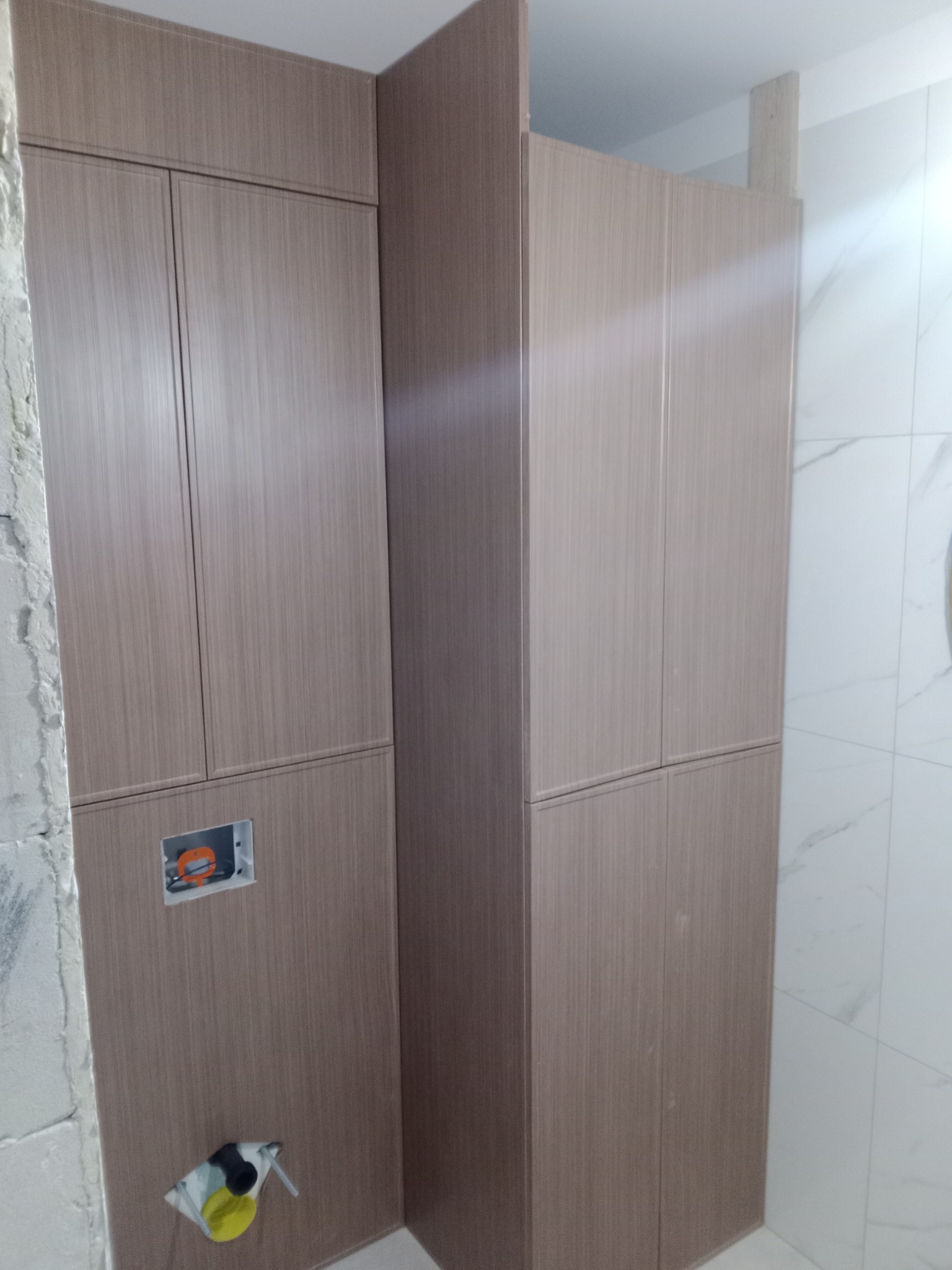 Kabinowa zabudowa z brązowych paneli w łazience, widoczne elementy instalacji sanitarnej i fragment surowej ściany.