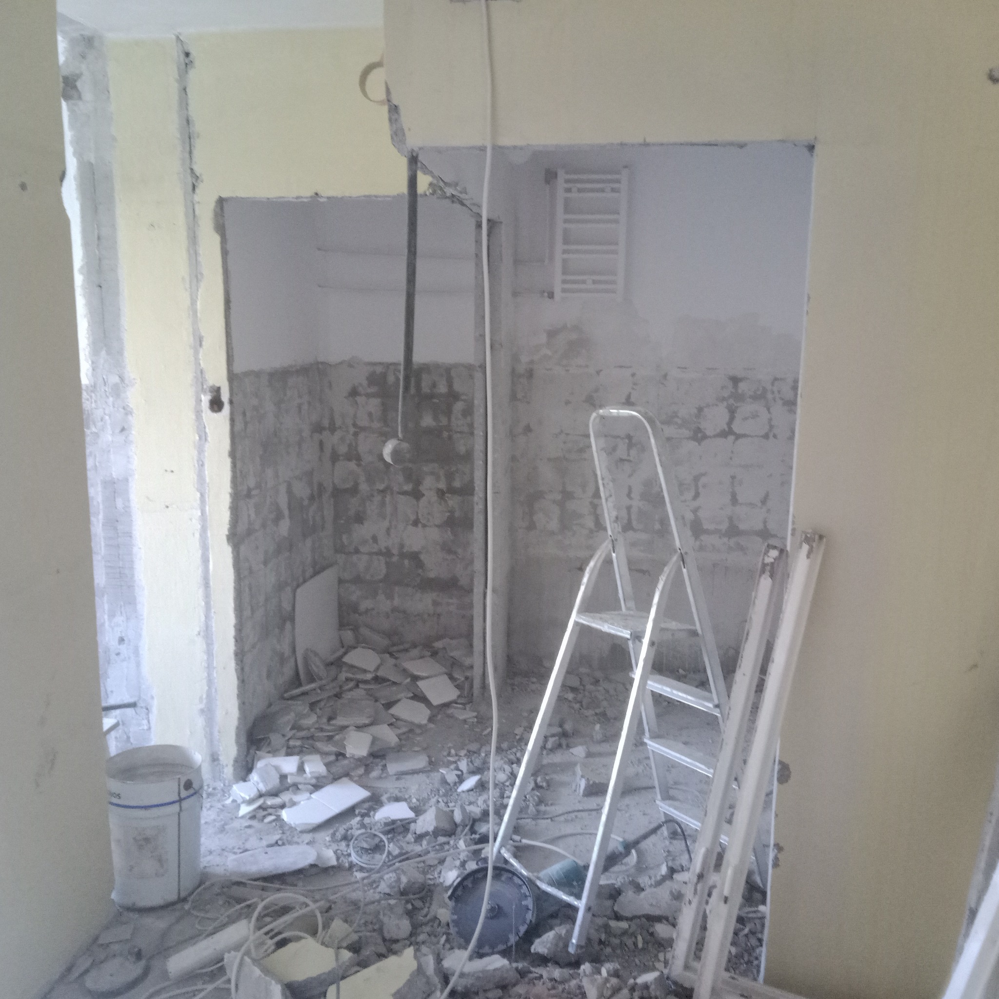 Wnętrze łazienki w trakcie demontażu płytek, widoczne resztki gruzu, drabina aluminiowa i wiadro po farbie.