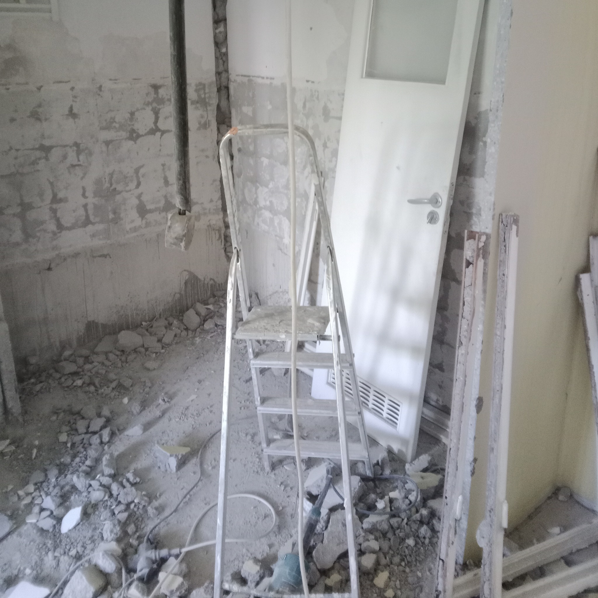 Wnętrze w trakcie demontażu: pobojowisko z gruzem, drabina aluminiowa, ościeżnica i drzwi z szybą, widoczne przewody i narzędzia.